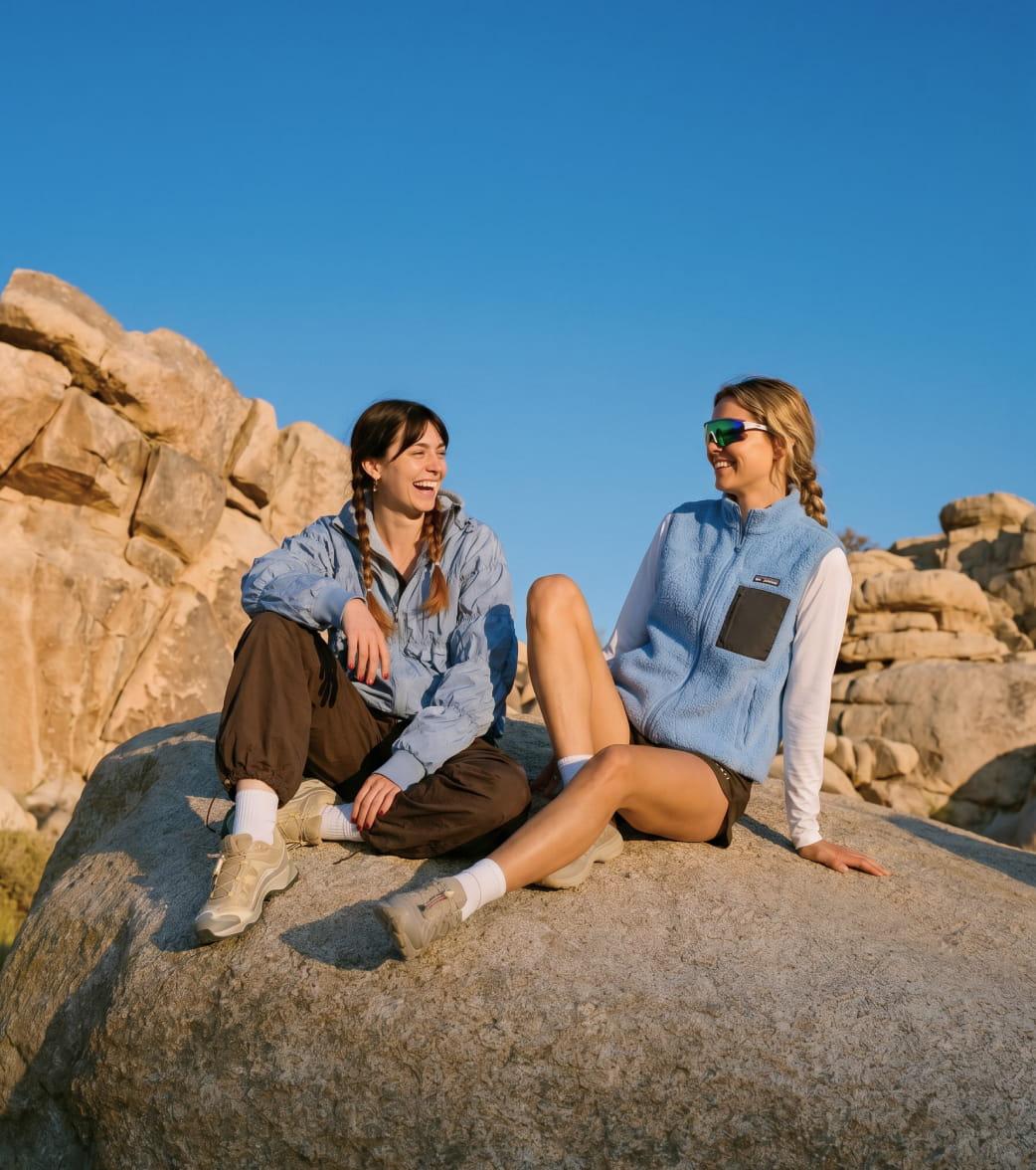 Two women sitting on a large rock in a desert landscape, wearing light blue fleece and zip-up outerwear from The North Face under a clear blue sky.